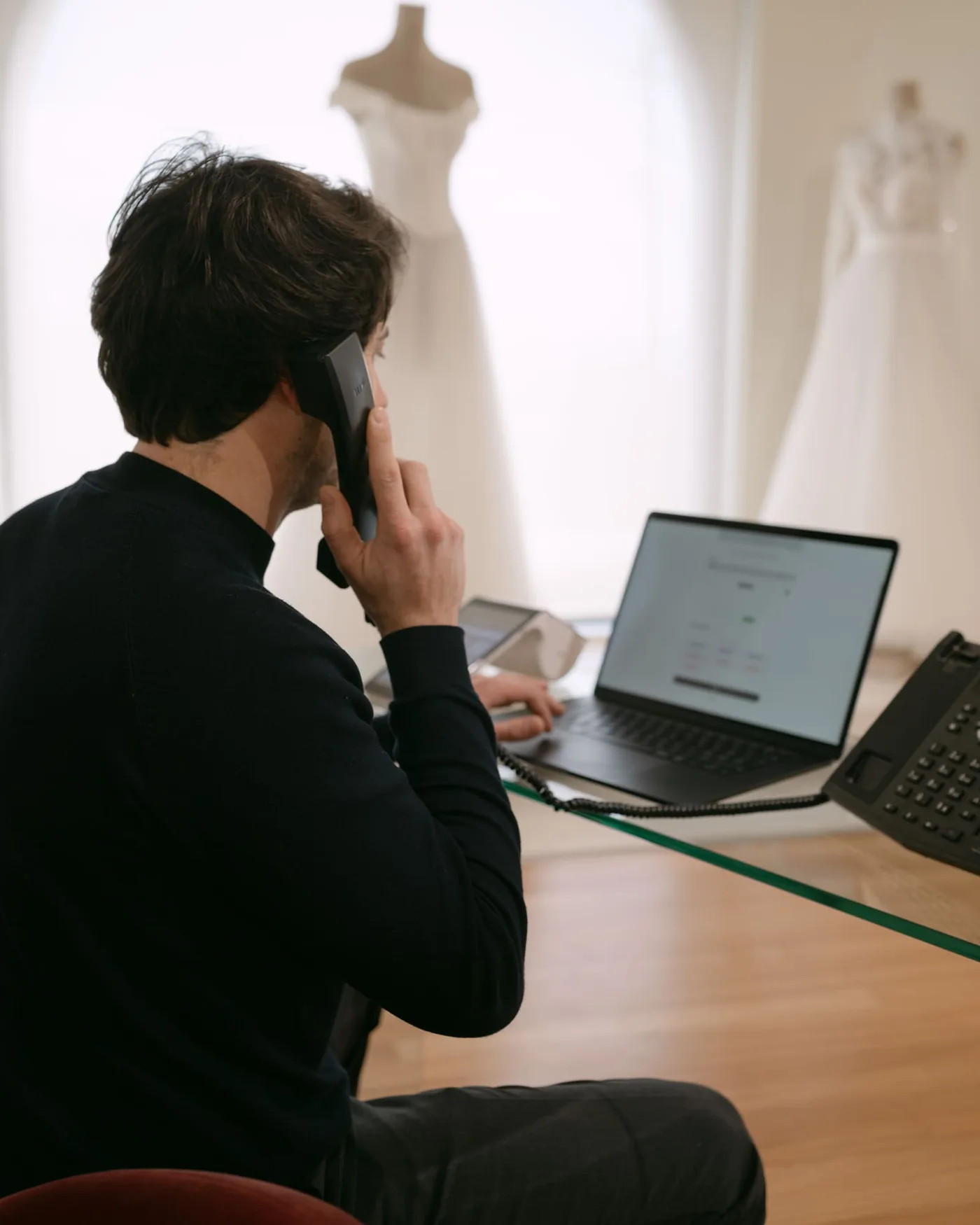 Dietro la scrivania di un atelier: una persona al telefono con il fisso, la mano sul portatile con il gestionale aperto; sullo sfondo, due abiti da sposa su manichino davanti alla vetrina.